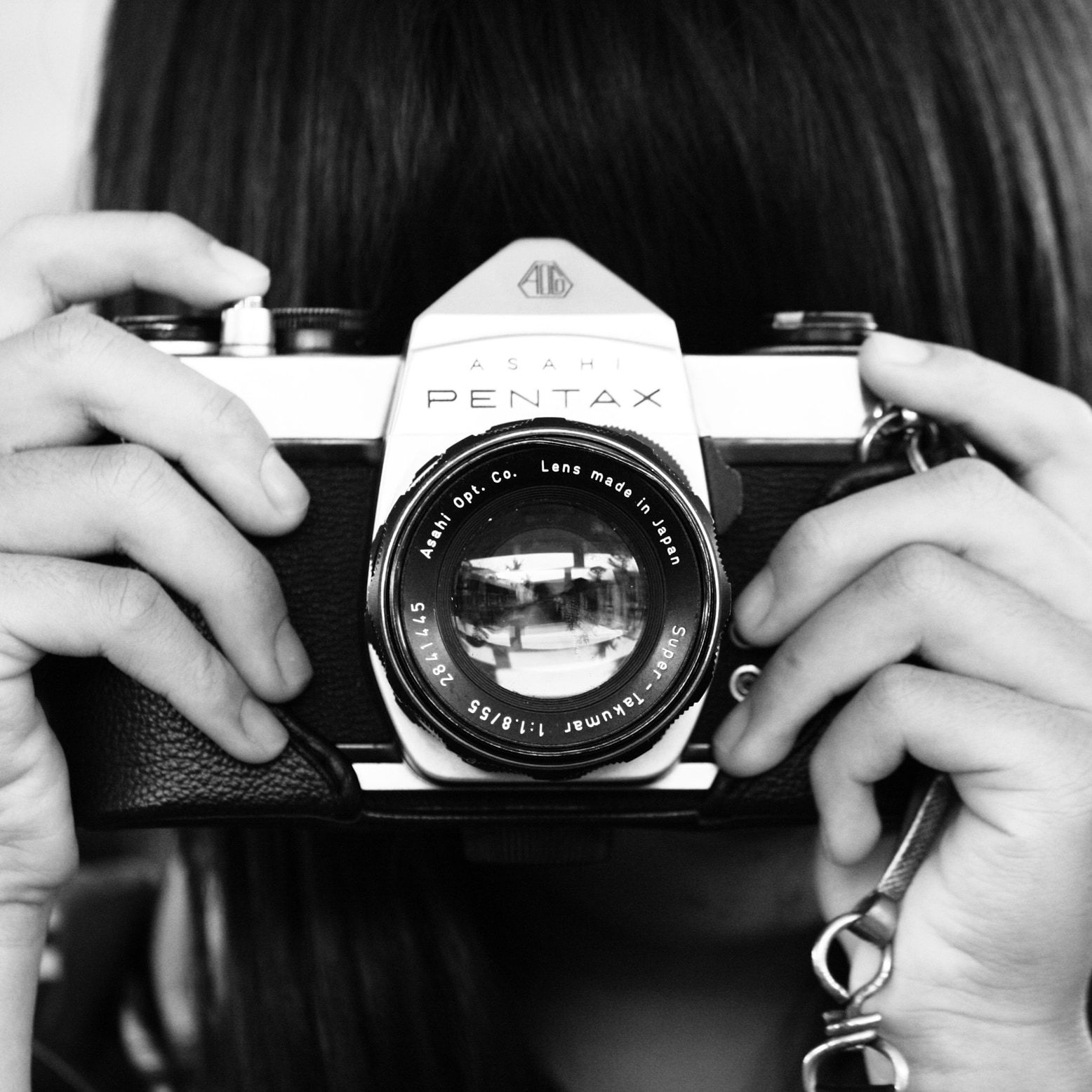 Unas manos sostienen una cámara analógica que apunta al frente. Fotografía en blanco y negro.